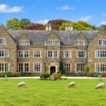 A large stone manor with a neatly trimmed lawn, several sheep grazing in front, and colourful trees and bushes surrounding the house on a clear, sunny day.