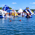 People enjoy an inflatable water park with slides and climbing structures on a lake, wearing life jackets under a clear blue sky. Some swim in the water whilst others play on the inflatables. Trees line the shore in the background.