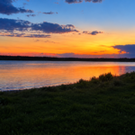 A vibrant sunset over a calm lake, with orange, yellow, and blue hues reflecting on the water, dark clouds above, and grassy shoreline in the foreground.