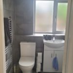 A modern bathroom with grey tiles, a white toilet, a white sink cupboard, a frosted window, a towel rail with a dark towel, and a black mat on the floor. Toilet paper and a toilet brush are next to the toilet.