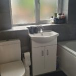 Modern bathroom with a toilet, white sink cupboard, and bathtub. Frosted window above the sink, with toiletries and cups on the windowsill. Dark floor mat and grey tiled walls complete the space.