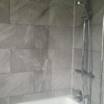 Modern bathroom with grey tiled walls, a white bath, a glass shower screen, and a chrome shower fitting with an overhead and handheld showerhead.