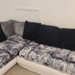 A cosy, L-shaped white sofa with black and grey cushions is placed in the corner of a white room. The sofa has textured, patterned throws in shades of grey and black.