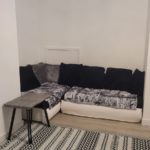 A cosy L-shaped sofa with dark and light cushions sits in a corner, next to a small grey table. A patterned black-and-white rug covers the light wood floor. The walls are plain white.