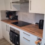 Modern kitchen with white cupboards, a wooden worktop, electric hob, oven, kettle, and utensils. There are kitchen roll on the top shelf and an open drawer with kitchen foil sticking out.