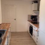 A modern galley kitchen with wooden worktops, white cupboards, a sink on the left, and an oven, hob, and appliances on the right. A closed door is at the end, and light streams in from the window.