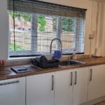 Modern kitchen with white cupboards, wooden worktop, large window with blinds, a sink, dish rack, soap bottle, kitchen roll, and a mobile on the worktop. Fence and greenery are visible outside the window.