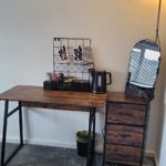 A tidy wooden desk with a kettle, mugs, snacks, and decor sits beside a small drawer unit and a floor lamp. A rubbish bin is underneath, and a small mirror hangs on the side. The carpet is grey.