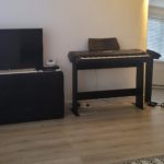 A black digital piano on a stand sits next to a TV on a low cabinet in a modern room with light wood flooring and a patterned rug.