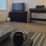 A black mug with a drink sits on a table in a modern living room with a rug, keyboard, telly, and a clock leant against a wall.