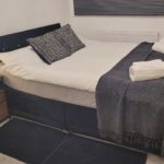 A neatly made bed with a dark blanket, two rolled white towels, and patterned cushions. A bedside table with a lamp and drawers is next to the bed. The room has grey carpet and closed blinds.