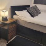 A neatly made bed with black and white pillows sits next to a wooden bedside table with two drawers and a modern lamp. The room has neutral colours and a cosy atmosphere.