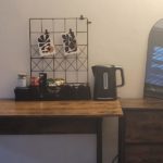 A wooden desk holds a wire grid with art, snacks, a kettle, and cups. A mirror sits on a matching drawer unit to the right. The wall and lighting are neutral.