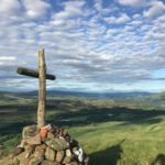A wooden cross stands on a mound of rocks atop a grassy hill, overlooking a vast, sunlit landscape of fields, hills, and distant water under a sky filled with scattered clouds.