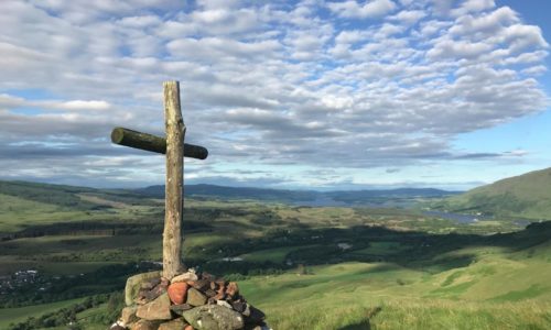 A wooden cross stands on a mound of rocks atop a grassy hill, overlooking a vast, sunlit landscape of fields, hills, and distant water under a sky filled with scattered clouds.