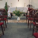 A small chapel with wooden chairs, a white-clothed altar, candles, a crucifix on the wall, and a vase of yellow flowers on a rug in the aisle.