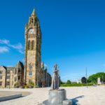 A tall stone clock tower stands beside historic buildings under a blue sky in Manchester. In the foreground, a statue of a woman holding a book is placed on a circular plinth in a spacious, paved square—an inviting spot often admired by visitors staying at the nearby Christian holiday home.