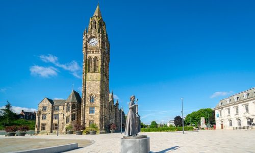 A tall stone clock tower stands beside historic buildings under a blue sky in Manchester. In the foreground, a statue of a woman holding a book is placed on a circular plinth in a spacious, paved square—an inviting spot often admired by visitors staying at the nearby Christian holiday home.
