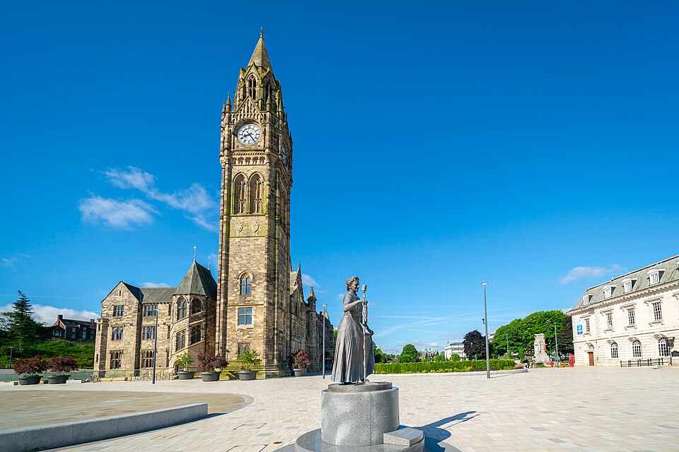 A tall stone clock tower stands beside historic buildings under a blue sky in Manchester. In the foreground, a statue of a woman holding a book is placed on a circular plinth in a spacious, paved square—an inviting spot often admired by visitors staying at the nearby Christian holiday home.