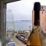 A view of a Tenby coastal street and the sea from a Christian holiday flat window, framed by a blurred wine glass and bottle in the foreground.