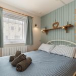 A bedroom in a Tenby holiday flat featuring a metal-framed bed with blue striped bedding, rolled brown towels, and a light blue panelled wall decorated with a wooden heart and four framed pictures.