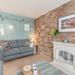 A cosy living room in a Tenby holiday flat featuring a grey sofa, a brick fireplace, light blue wood-panelled walls, and a coastal-themed print hanging above the mantelpiece.