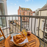 A basket of croissants, two glasses of juice, and a jar of jam on a small round wooden table at a Tenby holiday flat, overlooking a coastal town street.
