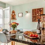 Dining table with a vase of flowers and place settings next to a kitchen island with wine, glasses, bread, and tomatoes in a Tenby holiday flat.