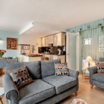 Open-plan living room and kitchen in a Tenby holiday flat, featuring two grey sofas with geometric print cushions, a dining area, and light green panelled walls.