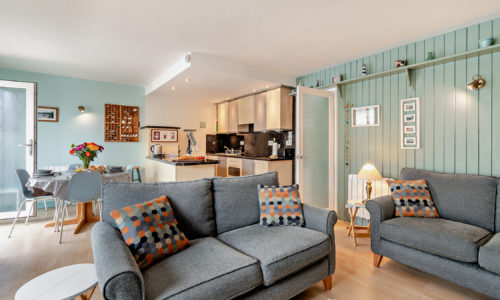Open-plan living room and kitchen in a Tenby holiday flat, featuring two grey sofas with geometric print cushions, a dining area, and light green panelled walls.