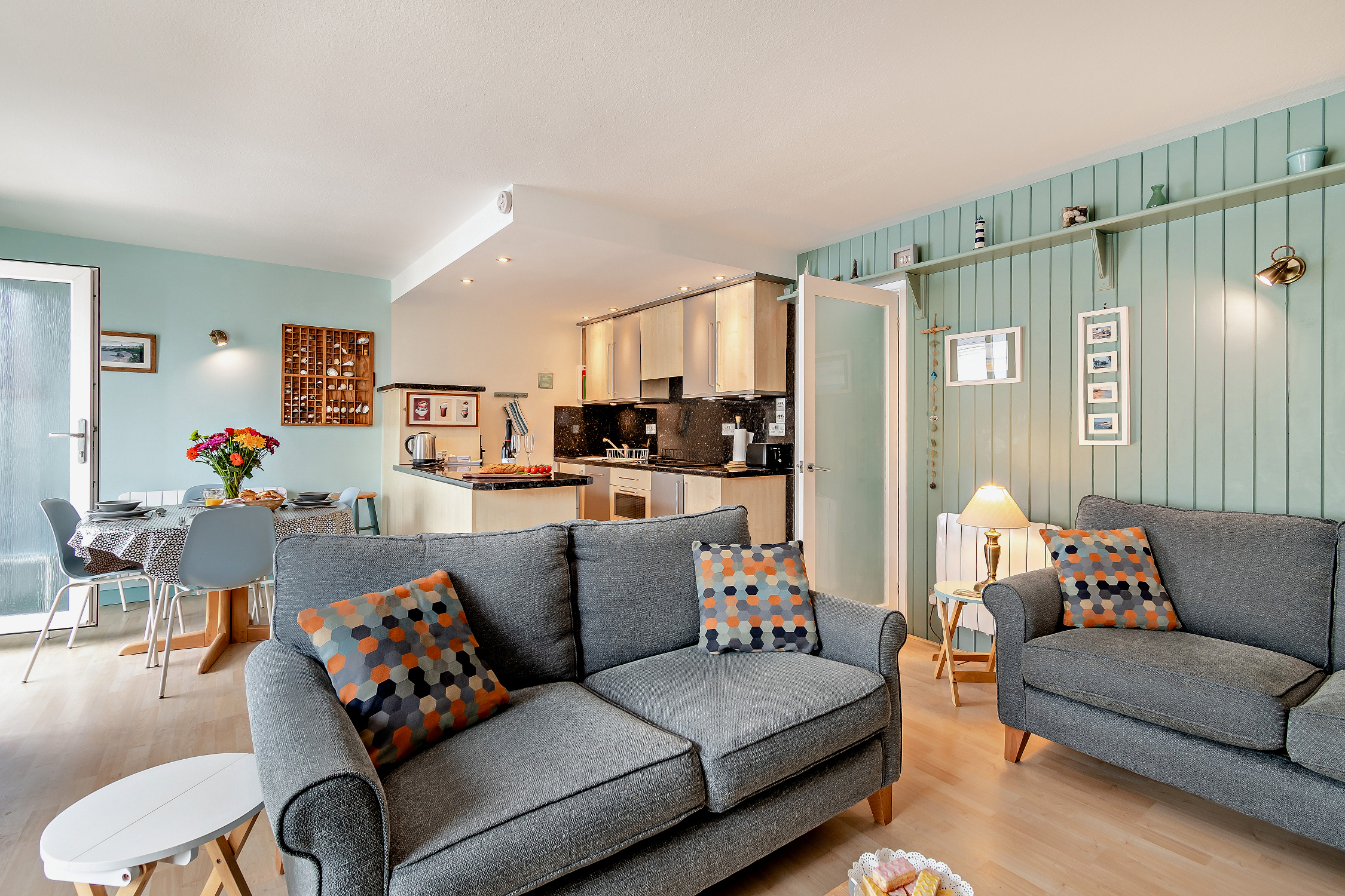 Open-plan living room and kitchen in a Tenby holiday flat, featuring two grey sofas with geometric print cushions, a dining area, and light green panelled walls.
