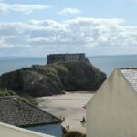A stone fort atop a rocky island overlooks a sandy beach and the sea, viewed from between the rooftops of a Tenby holiday flat.