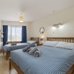 A bedroom in a Tenby holiday flat with a queen bed and a single bed, both featuring wooden frames and blue-striped bedding with rolled towels on top.